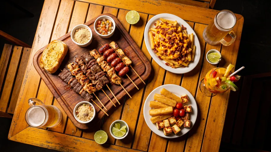 Mesa vista de cima com tábua de espetinhos, fritas com cheddar, mandioca com queijo coalho, chopp e drink.