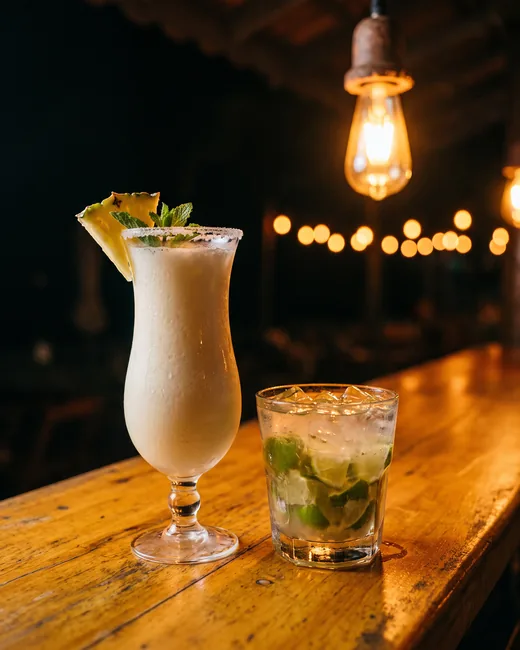 Piña colada em taça alta com abacaxi e hortelã ao lado de uma caipirinha, sobre balcão de madeira.