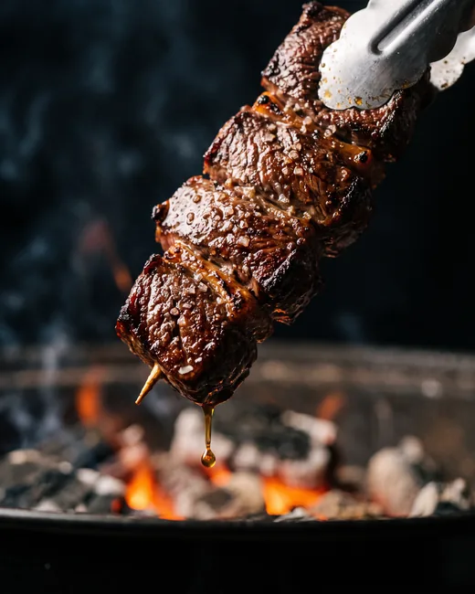 Espetinho de carne sendo retirado da brasa, com crosta caramelizada e sal grosso.