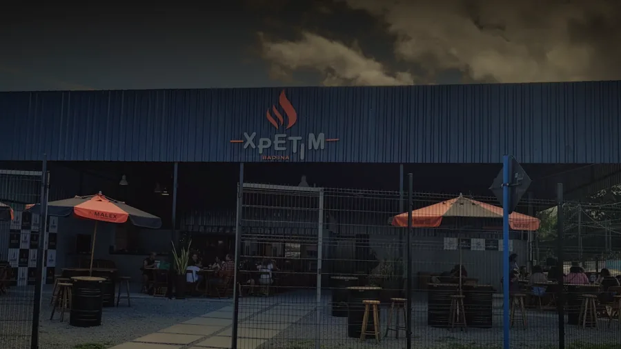 Fachada do Xpetim Maricá, galpão escuro com letreiro iluminado e mesas na área externa.