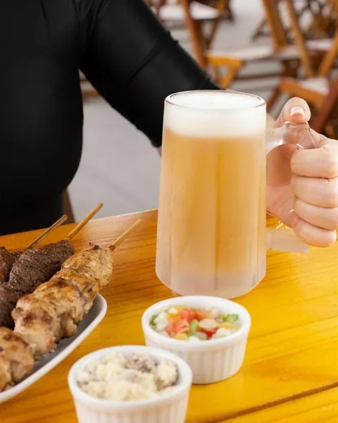 Caneca de chopp com colarinho, ao lado de espetinhos, farofa e vinagrete na mesa.