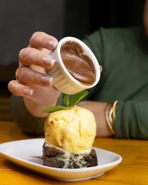 Brownie com bola de sorvete e calda de chocolate sendo despejada por cima.
