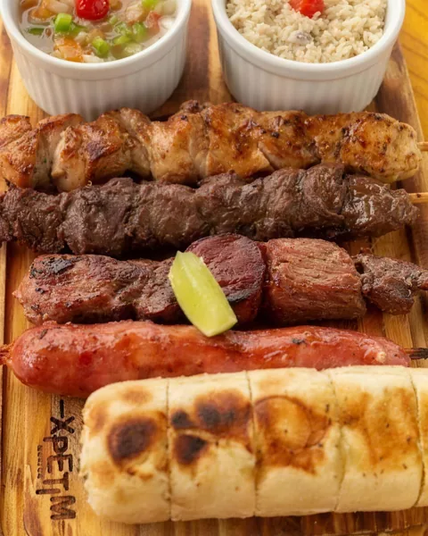 Tábua com espetinhos de frango, carne e linguiça, pão de alho, farofa e vinagrete.